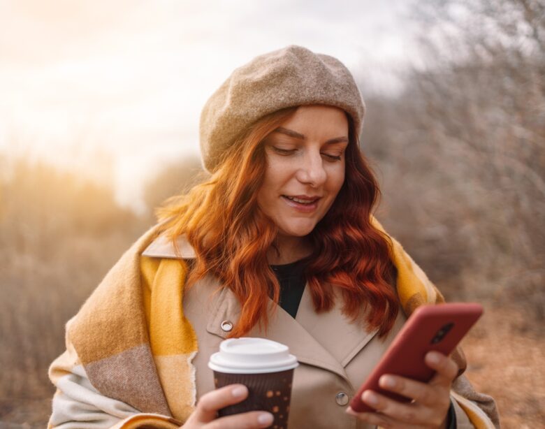Beautiful young woman using smartphone and drinking coffee takeaway at city park