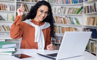 Delighted university student making eureka gesture while sitting by table with pc on background of