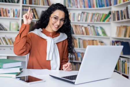 Delighted university student making eureka gesture while sitting by table with pc on background of